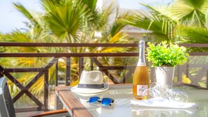 Een tafel met een champagnefles, een hoed, zonnebril en een bloempot op een balkon, met palmbomen op de achtergrond.