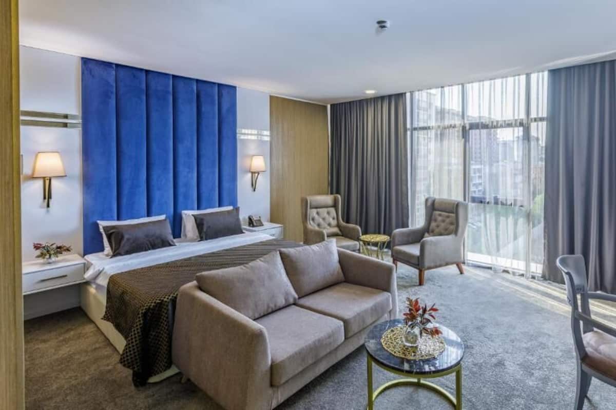 A hotel room with a large bed, two armchairs, a sofa, a small round table, and a view of the city through large windows.
