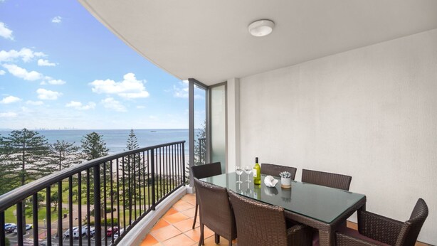 A balcony with a glass table and wicker chairs, overlooking a beach and city skyline. - Calypso Towers 901 - Coolangatta Beachfront - Min. 3 night stays! (Coolangatta)