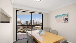 A room with a dining table, chairs, a view of the city through large windows, and a beach scene painting on the wall. - Calypso Towers 901 - Coolangatta Beachfront - Min. 3 night stays! (Coolangatta)