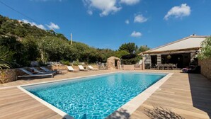En rektangulær swimmingpool med en træterrasse hele vejen rundt, en stenskulptur og en bygning med et tag af fliser i baggrunden.
