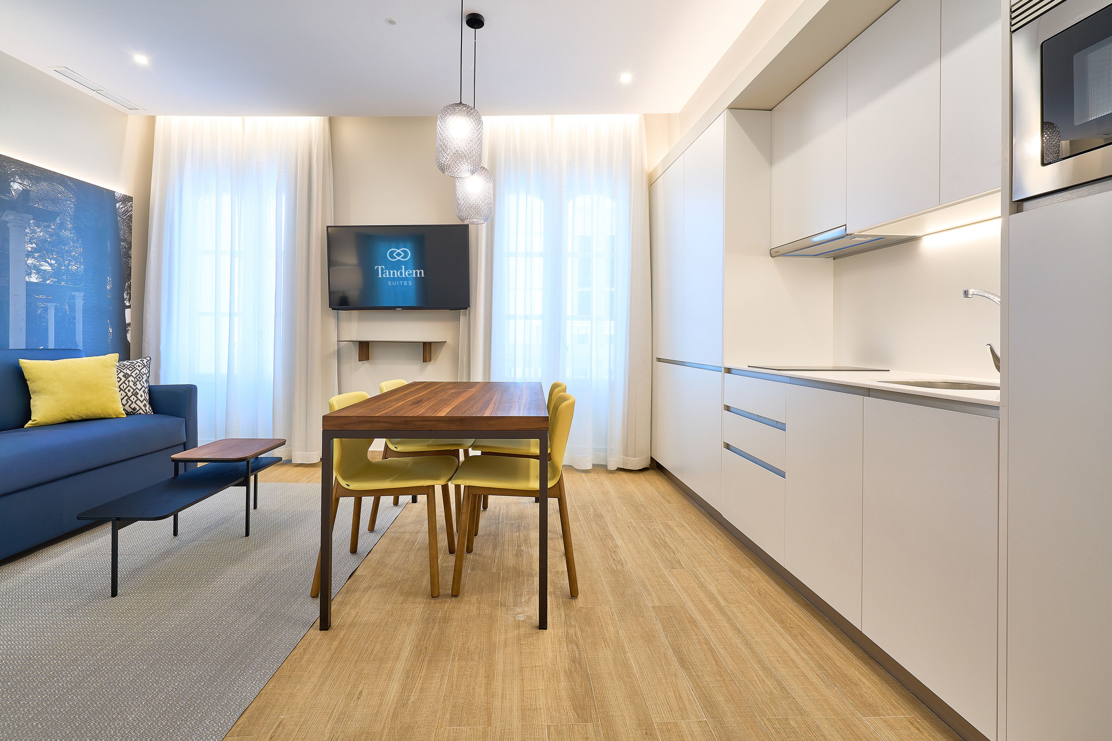 A modern living room with a blue sofa, a wooden dining table, and yellow chairs. A flat-screen TV is mounted on the wall above the sofa.