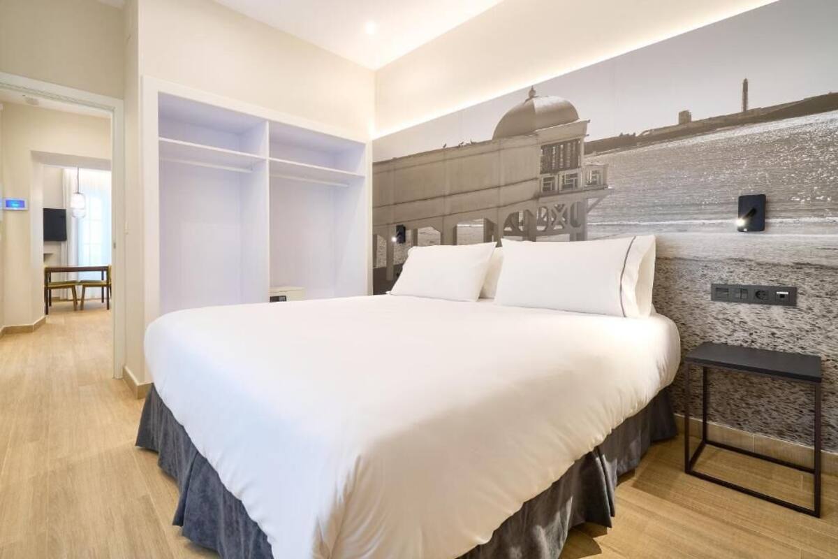 A hotel room with a large bed, a wardrobe, a small table, and a wall-mounted control panel.