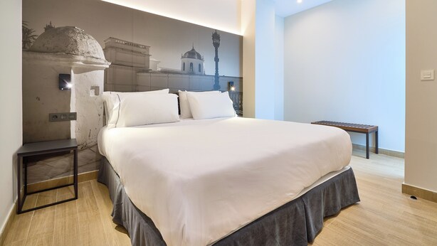 A neatly made bed with a dark skirt, a small bedside table, and a wall-mounted lamp. - Tándem Pópulo (Cádiz)