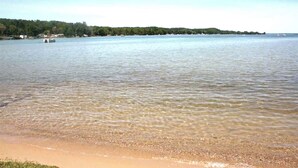 En strand med klart, lavt vand, en fjerntliggende kystlinje med træer, og en lille båd i vandet.