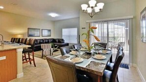 Uma área de refeições com uma mesa posta para quatro, um bar com banquetas, um sofá e uma janela grande com persianas.