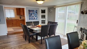 Un comedor con mesa de madera y sillas negras, una cocina con armarios de madera y un ventanal con persianas.