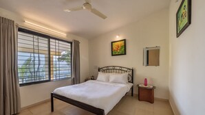 A bedroom with a bed, a ceiling fan, a window with curtains, a bedside table, and a wall-mounted picture. - CosmicStays Countryside Panorama-Lakeview Bungalow (Paud)