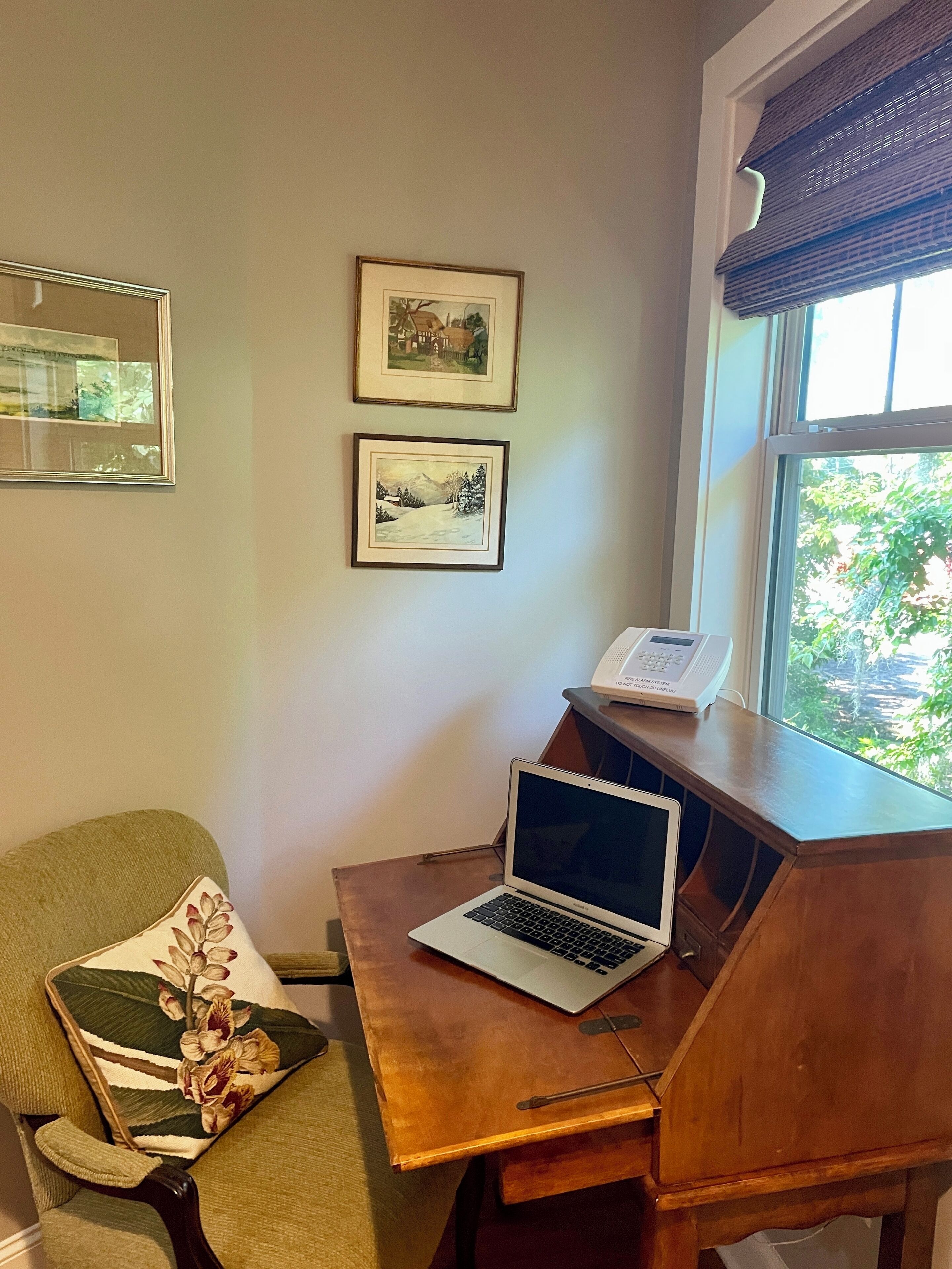 A room with a wooden desk, a laptop, a chair with a floral pillow, and two framed pictures on the wall.