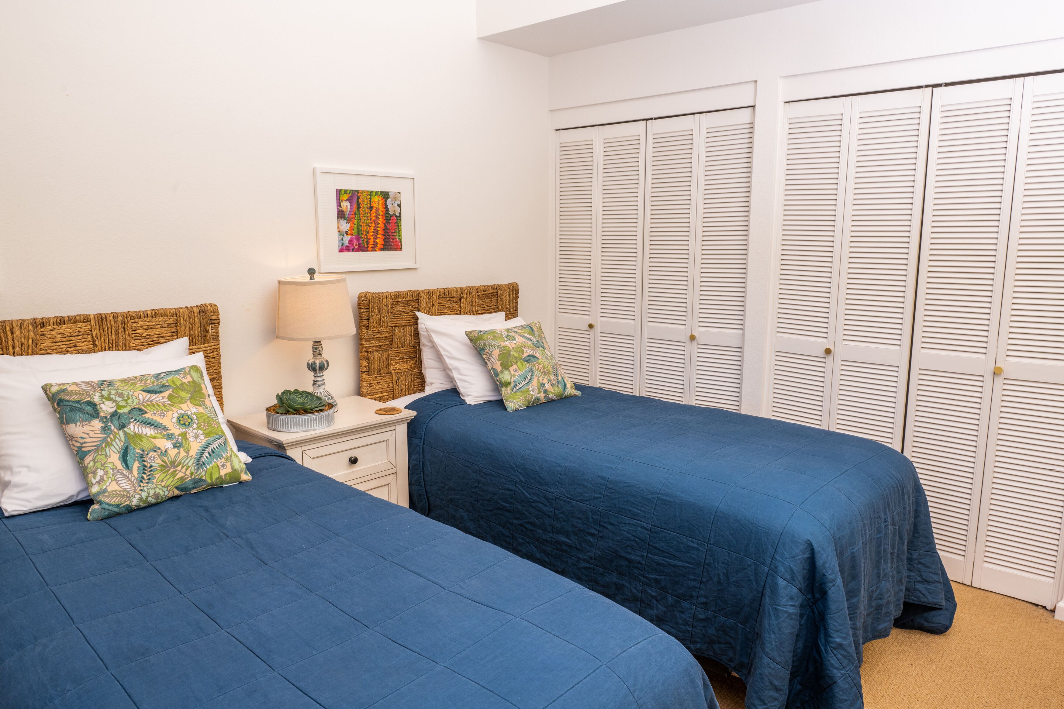 A bedroom with two beds, white walls, a framed picture, a nightstand, and a closet with louvered doors.