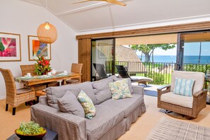 A living room with a grey sofa, wicker chairs, a dining table with chairs, and a view of the ocean through large sliding doors.