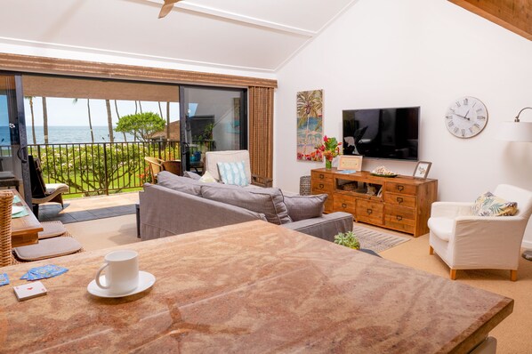 A spacious living area with a large sliding glass door leading to a balcony with a view of the ocean. There is a comfortable seating area with a sofa and a white armchair, a wooden cabinet with a flat-screen TV, and a dining table with a coffee cup and playing cards.