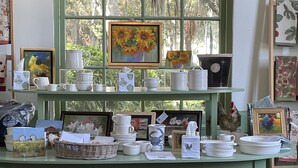 A room with a green wall and a window with a view of trees. There are shelves with various items like plates, cups, and paintings.