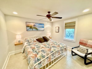 A bedroom with a bed, a ceiling fan, a window with a view, and a chair with a sign.
