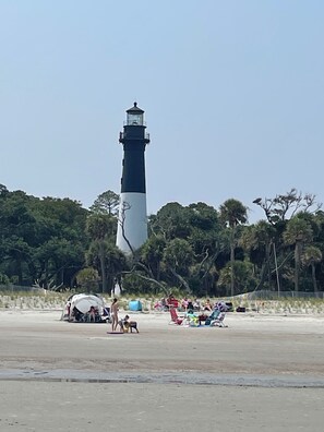 Un phare aux couleurs blanc et noir, entouré d’arbres et bordé d’une plage de sable.