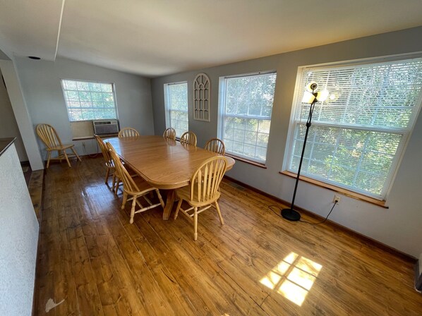 Une salle à manger avec une table et des chaises en bois, de grandes fenêtres équipées de stores et un lampadaire.
