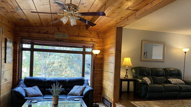 A cozy cabin interior with a blue leather sofa, a glass-top coffee table, a ceiling fan, and a large window offering a view of trees. - Beautiful Home in Glendo (Glendo)