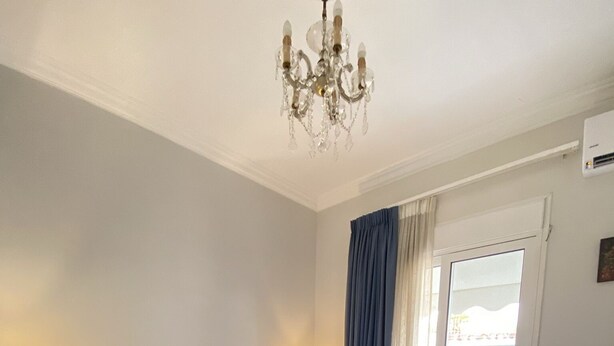 A neatly made bed with a blue bedspread and white pillows, a bedside lamp, a wooden nightstand, a chandelier, and a sliding glass door with curtains. - A Skip Away from the Acropolis Museum (Athens)
