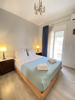 A neatly made bed with a blue bedspread and white pillows, a bedside lamp, a wooden nightstand, a chandelier, and a sliding glass door with curtains. - A Skip Away from the Acropolis Museum (Athens)