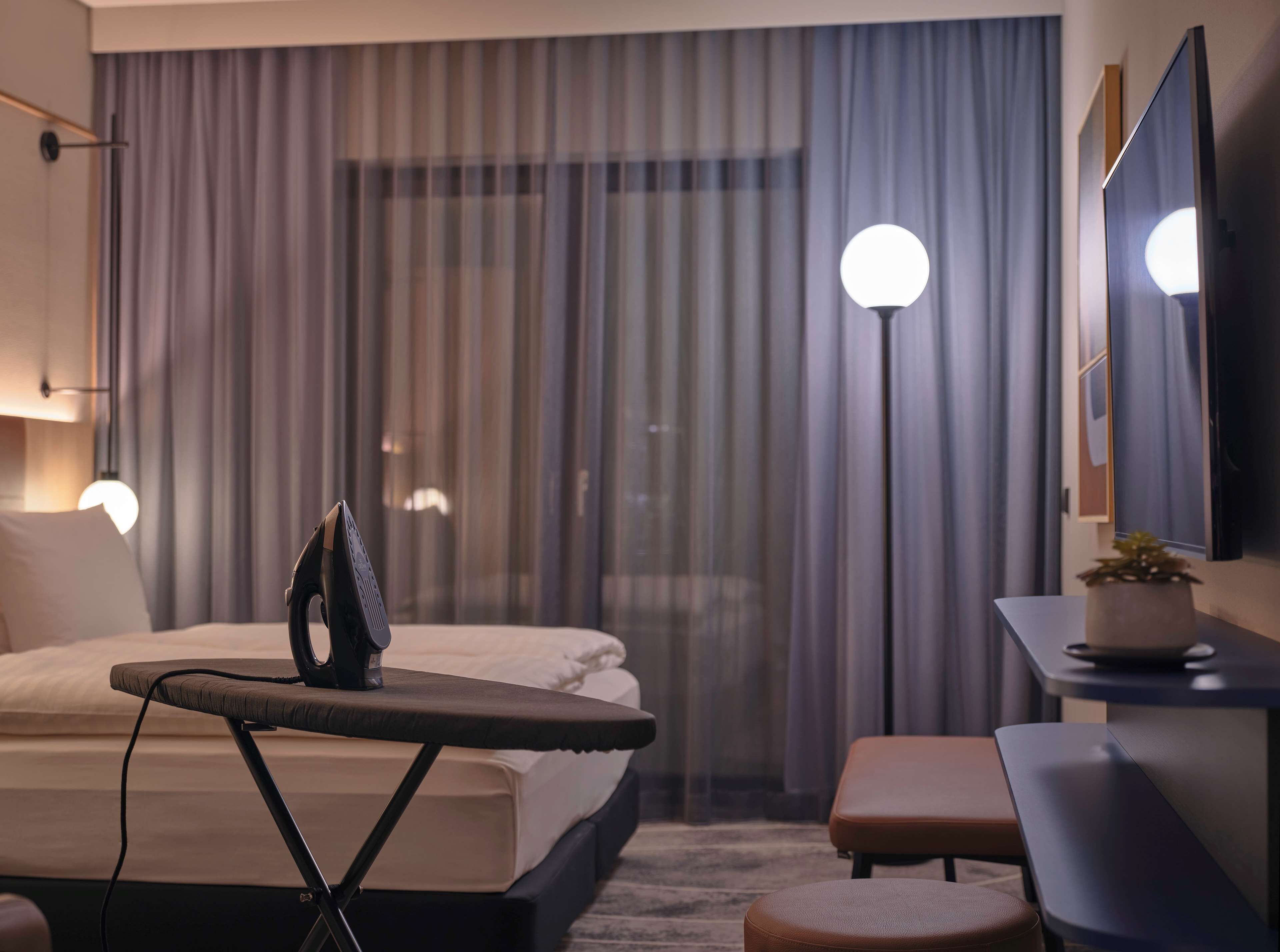 A modern hotel room with a neatly made bed, an ironing board, a small table with a plant, and a large mirror on the wall.