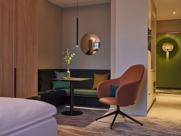 A modern hotel room with a green sofa, a round mirror, a small table with a vase of flowers, a brown armchair, and a bed.
