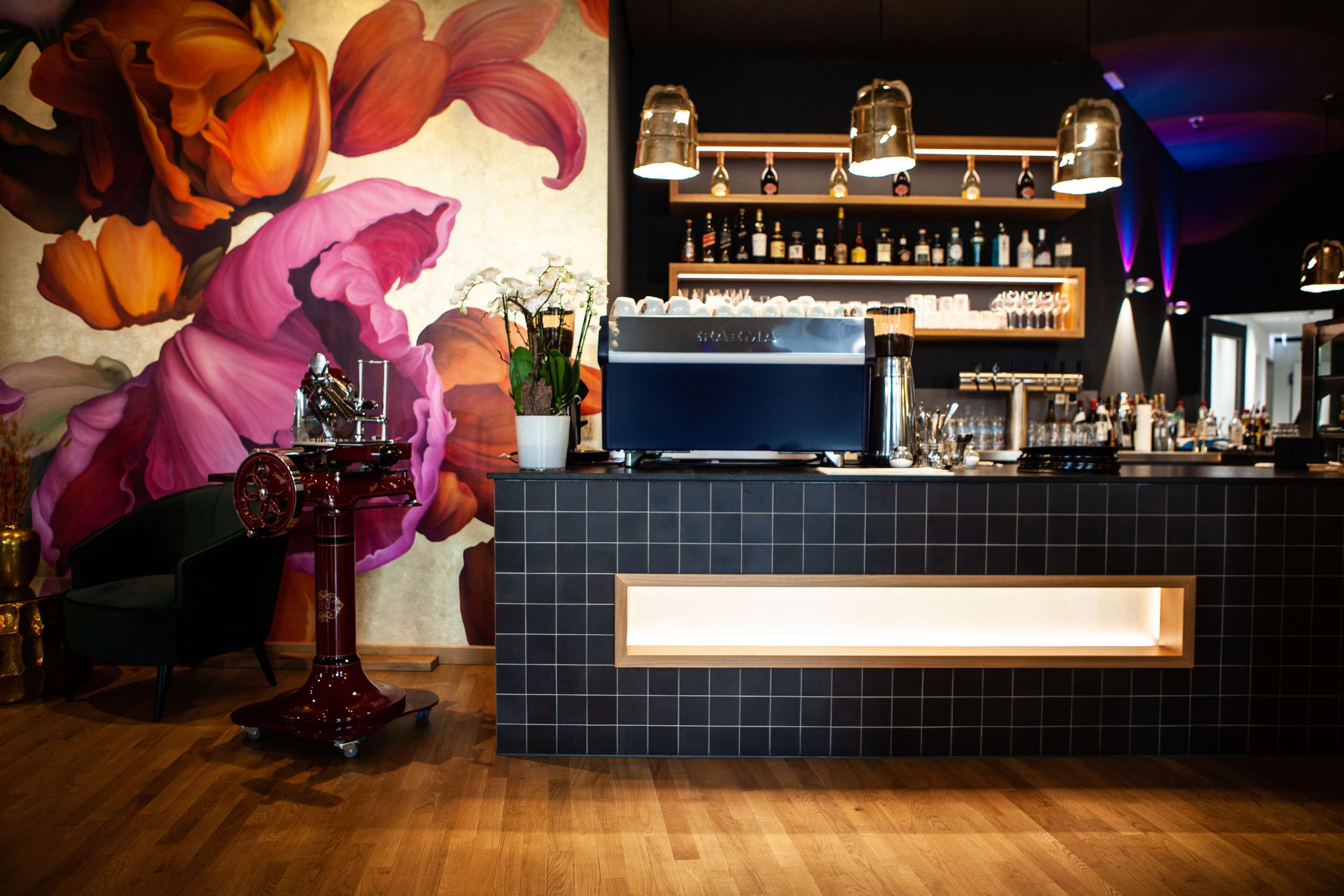 Uma área de bar com balcão revestido de azulejos escuros, uma máquina de café e prateleiras com garrafas.