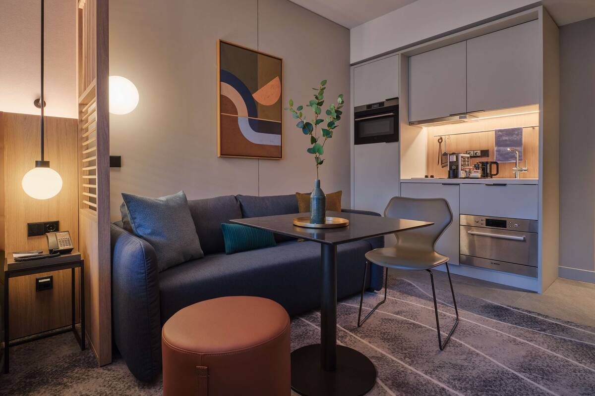 Ein modernes Wohnzimmer mit einer Couch, einem Couchtisch und einem kleinen runden Hocker. Es gibt eine Küchenzeile mit einer Mikrowelle und einer Kaffeemaschine.
