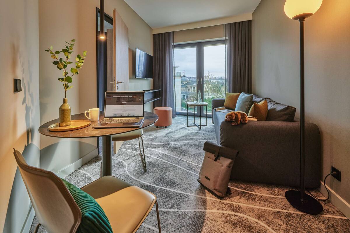 Ein modernes Hotelzimmer mit einem Laptop auf einem Schreibtisch, einem Sofa mit einer Katze, einer Pflanze und einem großen Fenster mit Blick auf die Stadt.