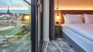 Hotellihuone, jossa on suuri ikkuna, josta avautuu näkymä kaupunkiin, siististi pedattu sänky, yöpöytä lampun kanssa ja matolla peitetty lattia.