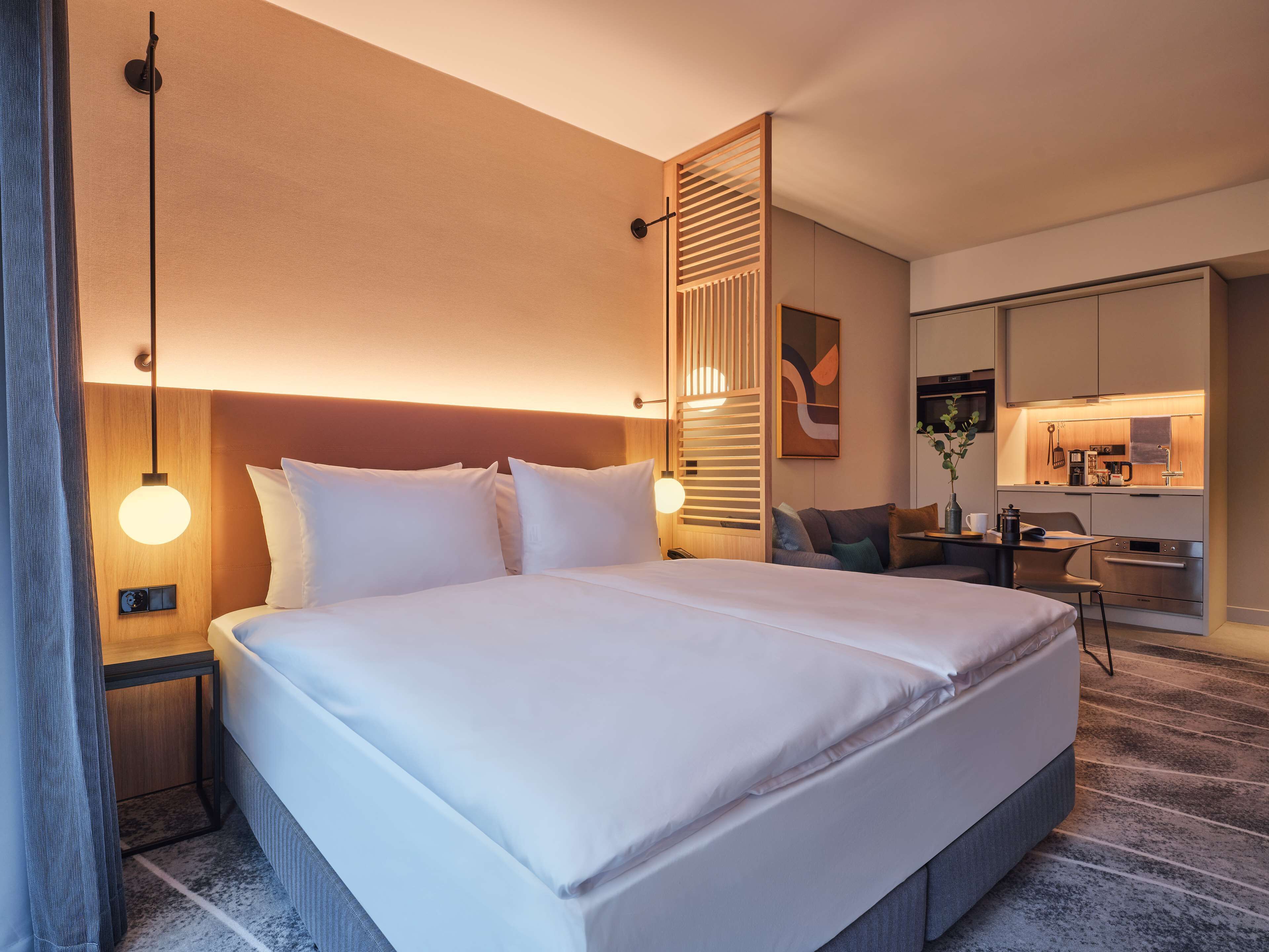 A modern hotel room with a large bed, bedside tables, a desk, and a kitchenette.