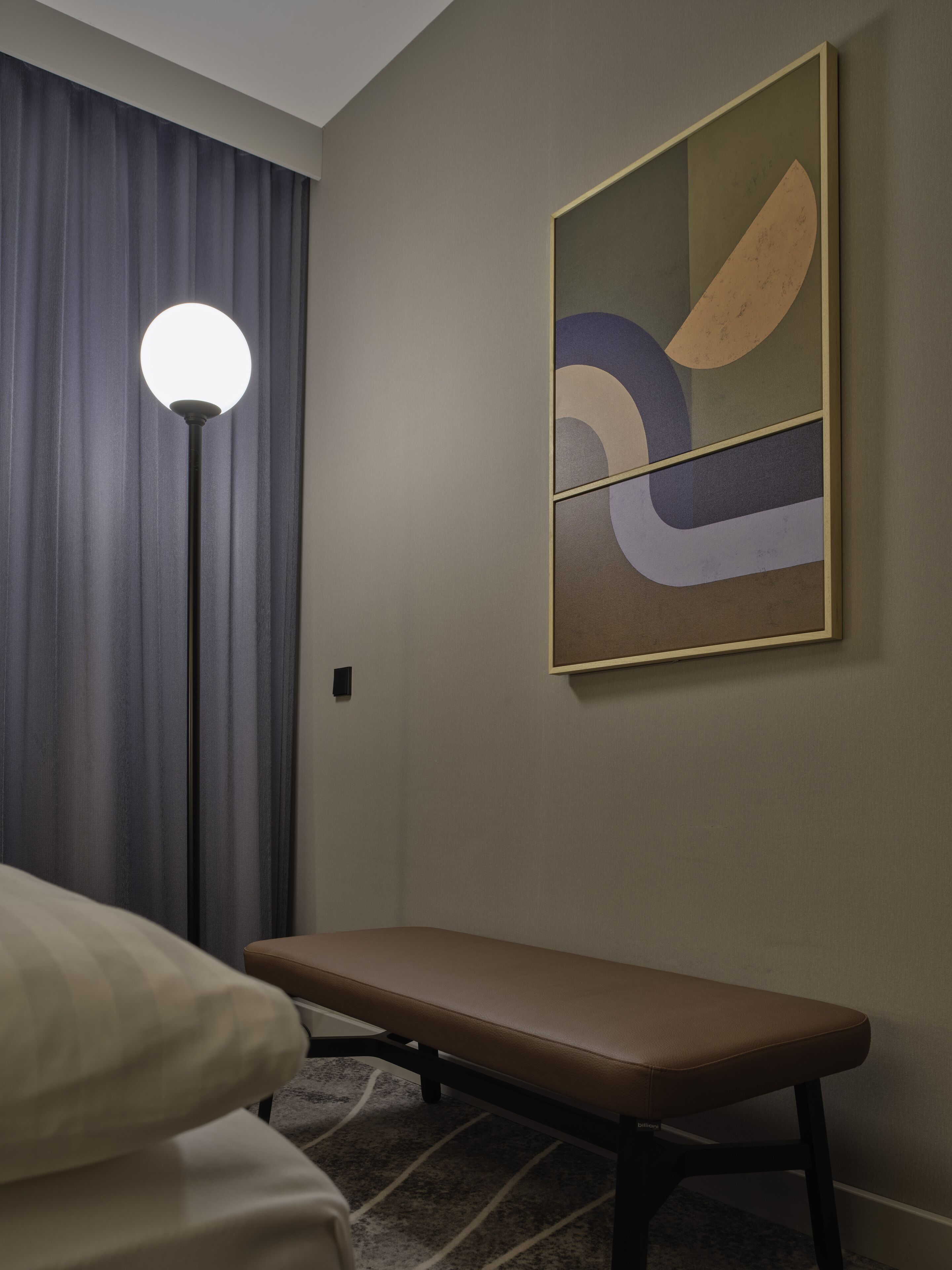 A hotel room with a bed, a bench, a floor lamp, and a wall-mounted abstract artwork.