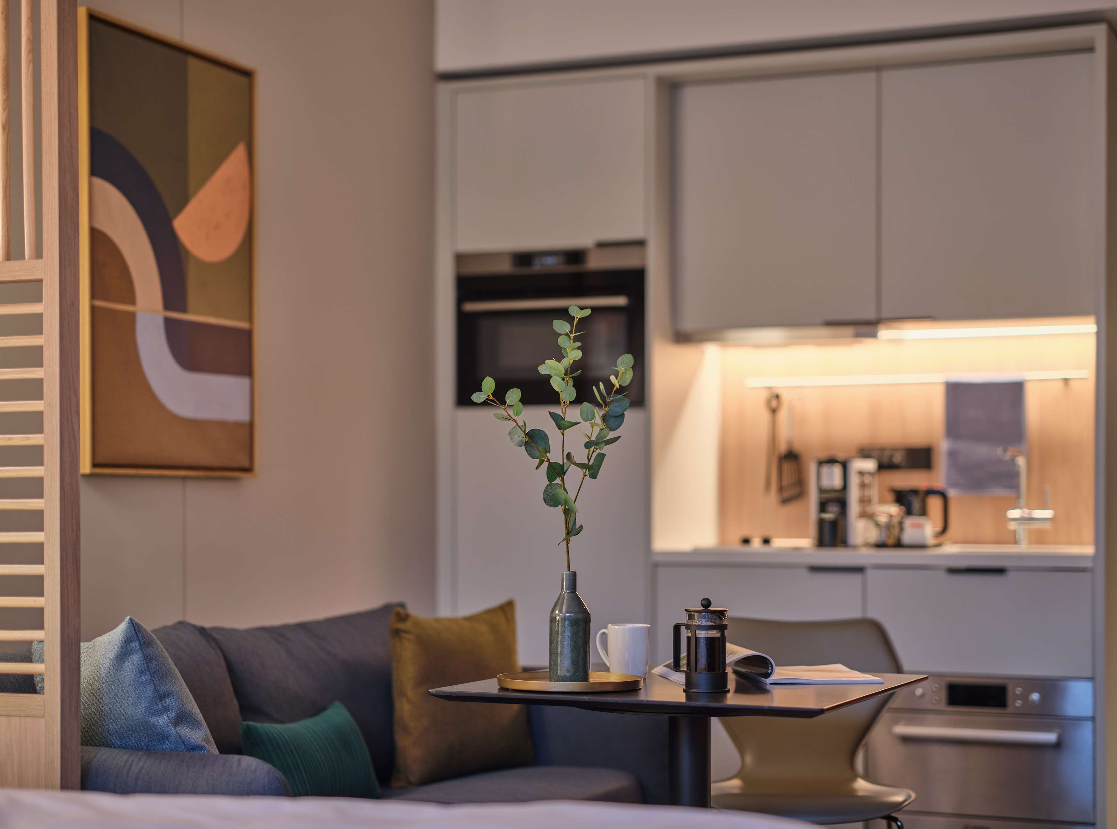 Una habitación de hotel moderna con un sofá, una mesita con un florero y follaje, una cafetera y una kitchenette al fondo.