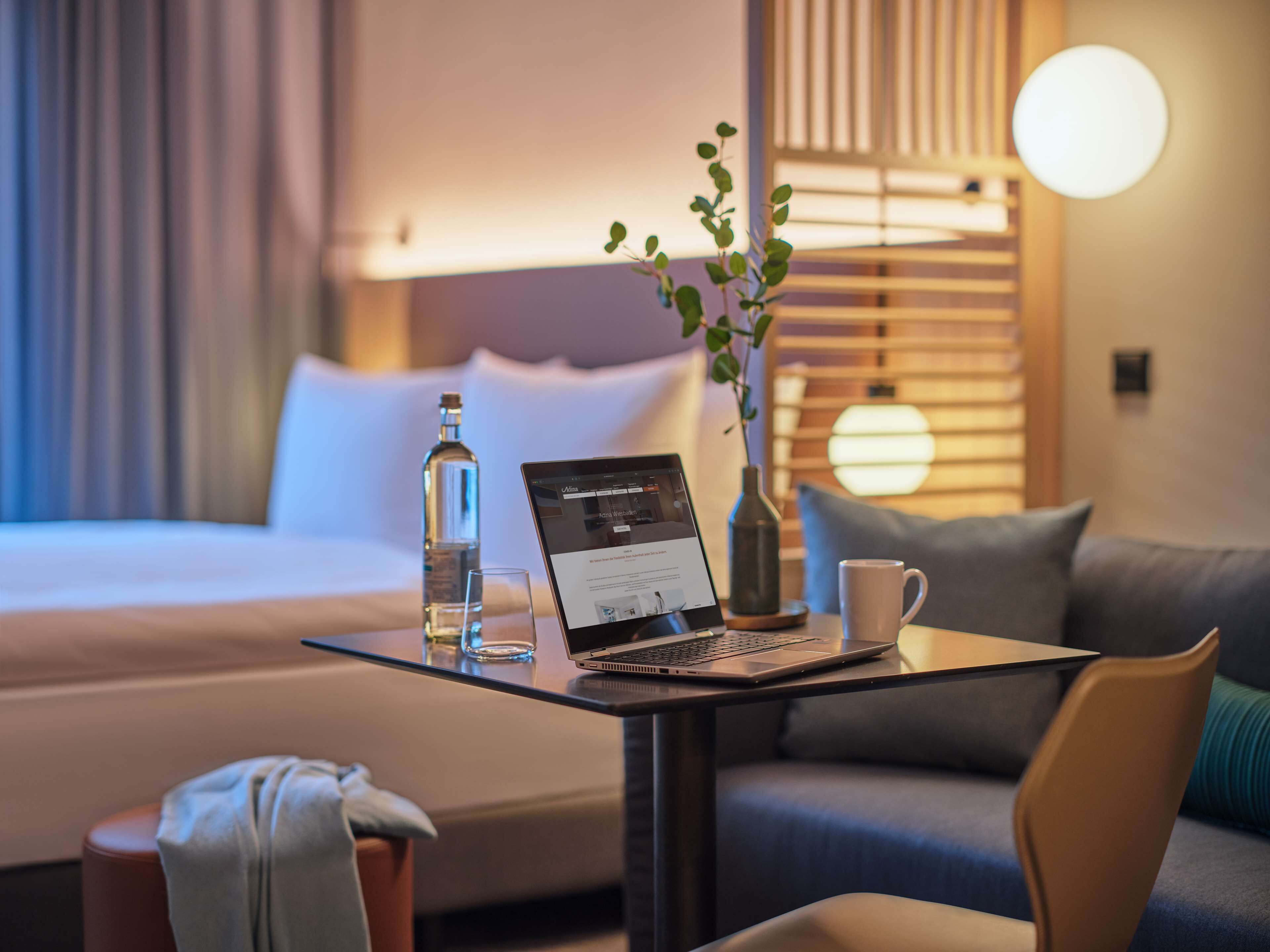 A hotel room with a bed, a laptop on a table, a bottle, a cup, and a vase with a plant.