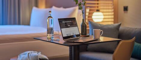 A hotel room with a bed, a laptop on a table, a bottle, a cup, and a vase with a plant.