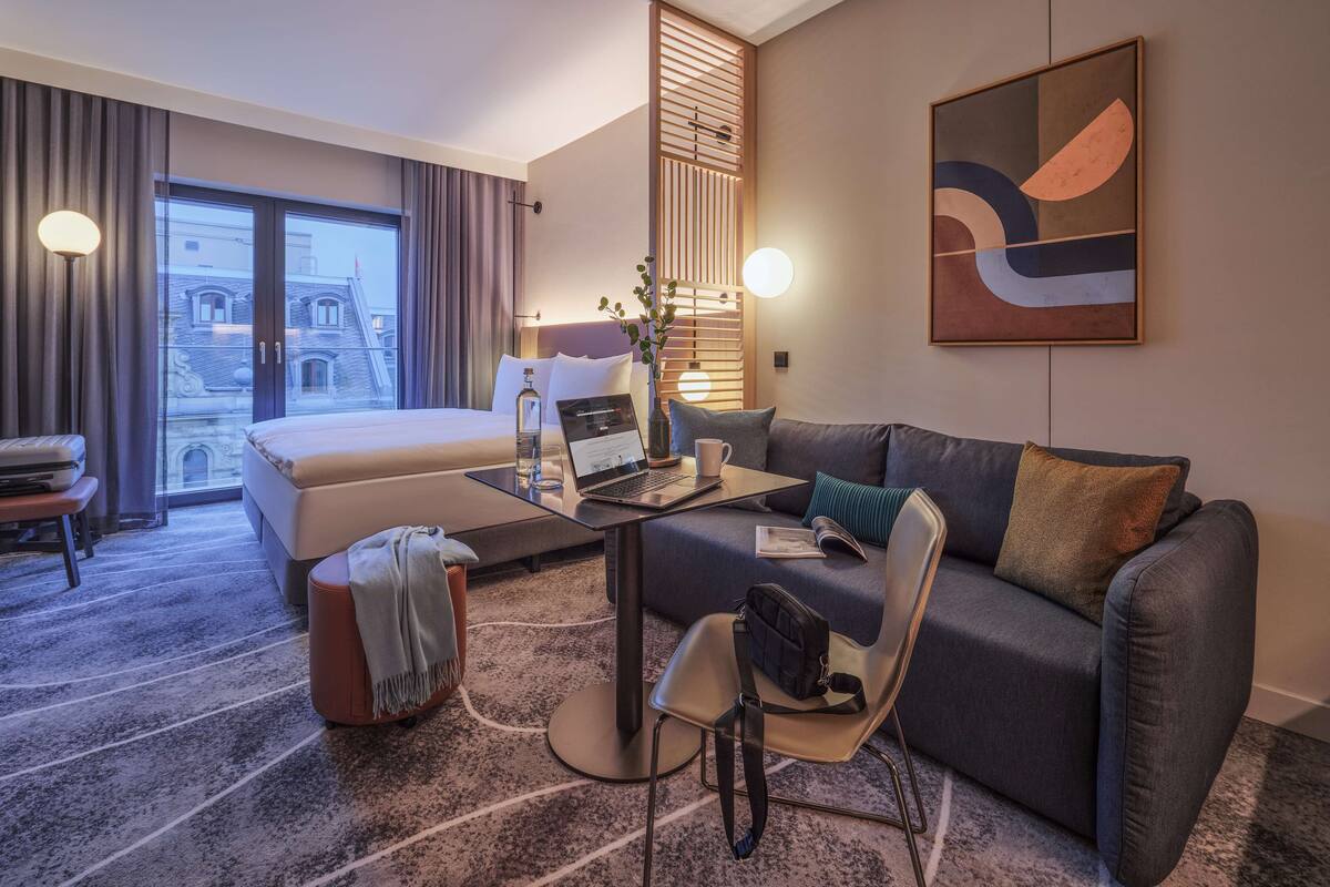 Ein modernes Hotelzimmer mit Bett, Sofa, Schreibtisch mit Laptop, Stuhl und Blick auf die Stadt.