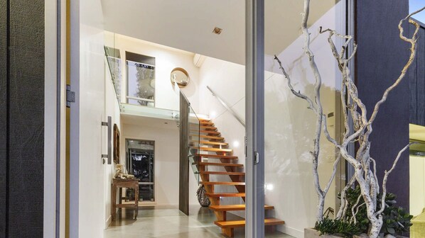 A modern interior with a wooden staircase leading to a loft area, a glass door entrance, and a minimalist decor with a white tree sculpture.