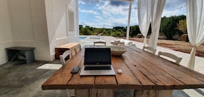 A wooden table with a laptop, a smartphone, and a white object, likely a speaker, in a covered outdoor area with a pool and greenery in the background.