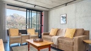 Ein Wohnzimmer mit einer Couch, zwei Sesseln, einem Couchtisch aus Holz und Blick auf Berge durch große Fenster.