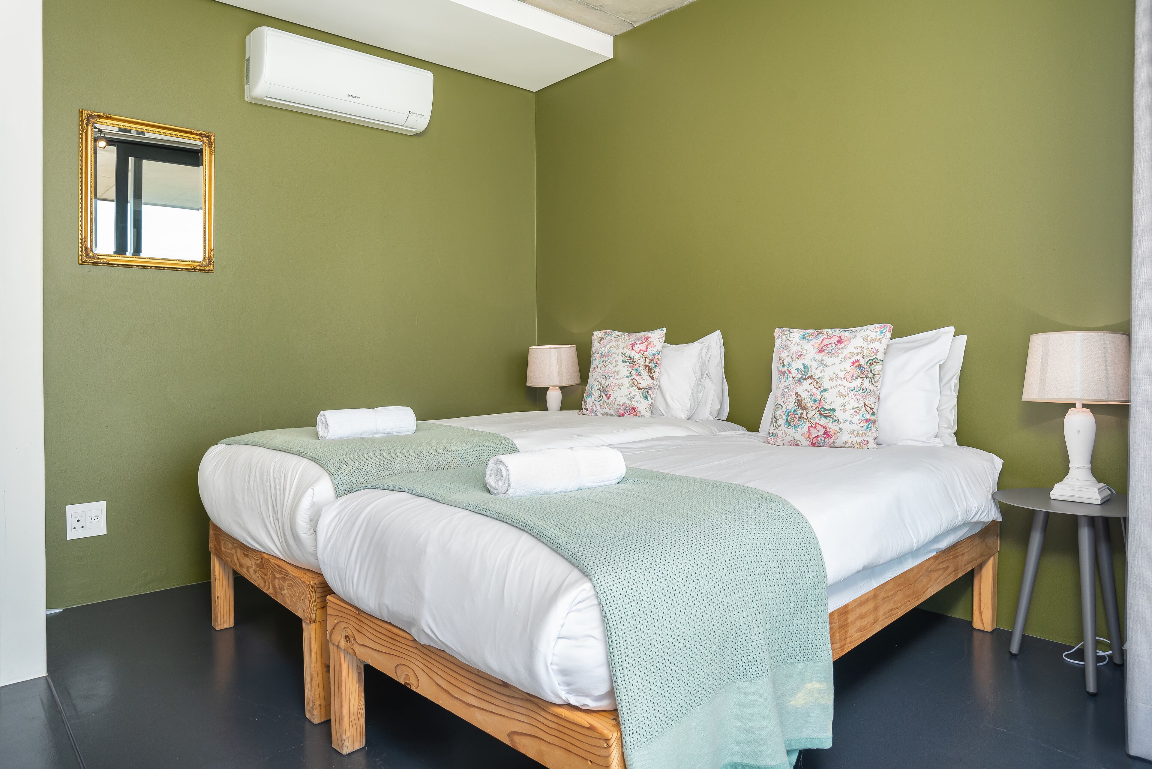 A double bed with white linens, a green throw blanket, and floral pillows. There is a bedside table with a lamp, a small mirror, and an air conditioning unit on the wall.