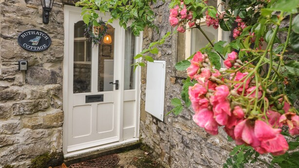 A stone cottage with a white door, a sign reading "Nuthatch Cottage", and a hanging light fixture. - Nuthatch Cottage (Skipton)
