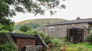 A stone house with a small window, a wooden door, and a garden area with a table and chairs. - Nuthatch Cottage (Skipton)