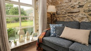 A cozy room with a stone wall, a window with a view of greenery, a grey sofa with cushions, and a table lamp. - Nuthatch Cottage (Skipton)