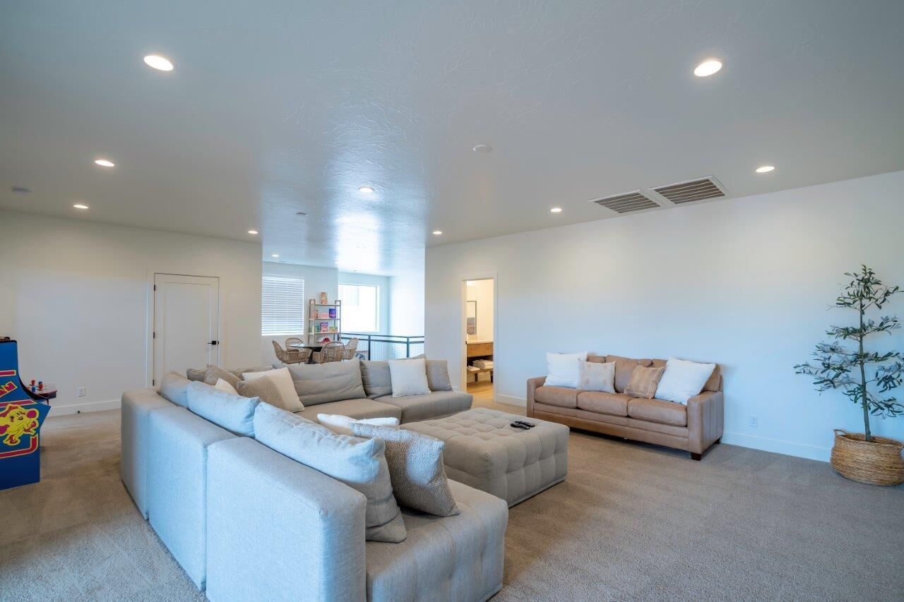 A spacious living room with a light-colored sectional sofa, a smaller sofa, and a coffee table. There is a potted plant and a small play area with toys visible.