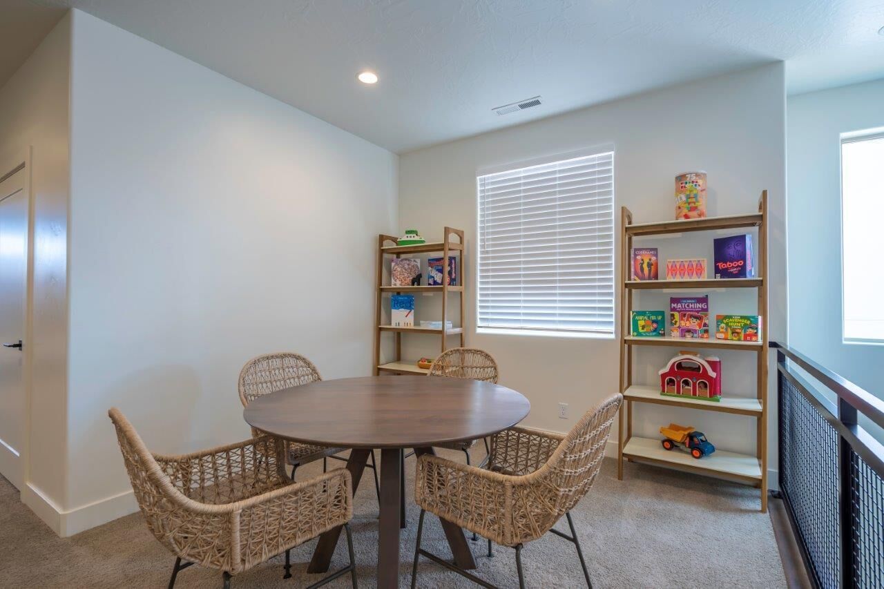 A room with a round table and wicker chairs, a bookshelf with children's toys, and a window with blinds.