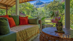 Ein gemütlicher Sitzbereich im Freien mit einer Couch und Kissen, einem kleinen Tisch mit Buddha-Statue und Blick auf üppiges Grün und Berge.