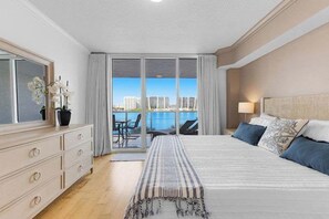 A hotel room with a large bed, a dresser, a view of the water, and a balcony with chairs. - Le Marina Bay 409  	STR-03586 (Sunny Isles Beach)