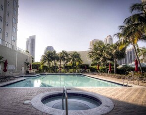 A swimming pool with a hot tub, surrounded by palm trees and red umbrellas, with tall buildings in the background. - Le Marina Bay 409  	STR-03586 (Sunny Isles Beach)