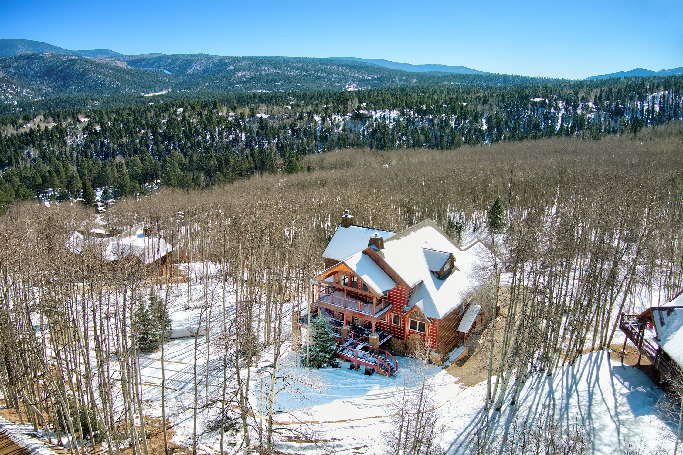 A log cabin with a large deck, surrounded by snow-covered ground and trees, with mountains in the background.