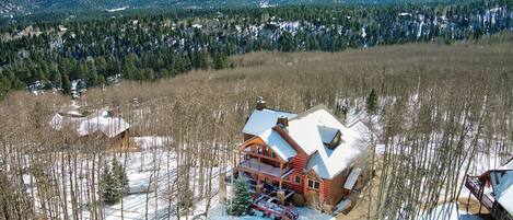 A log cabin with a large deck, surrounded by snow-covered ground and trees, with mountains in the background.