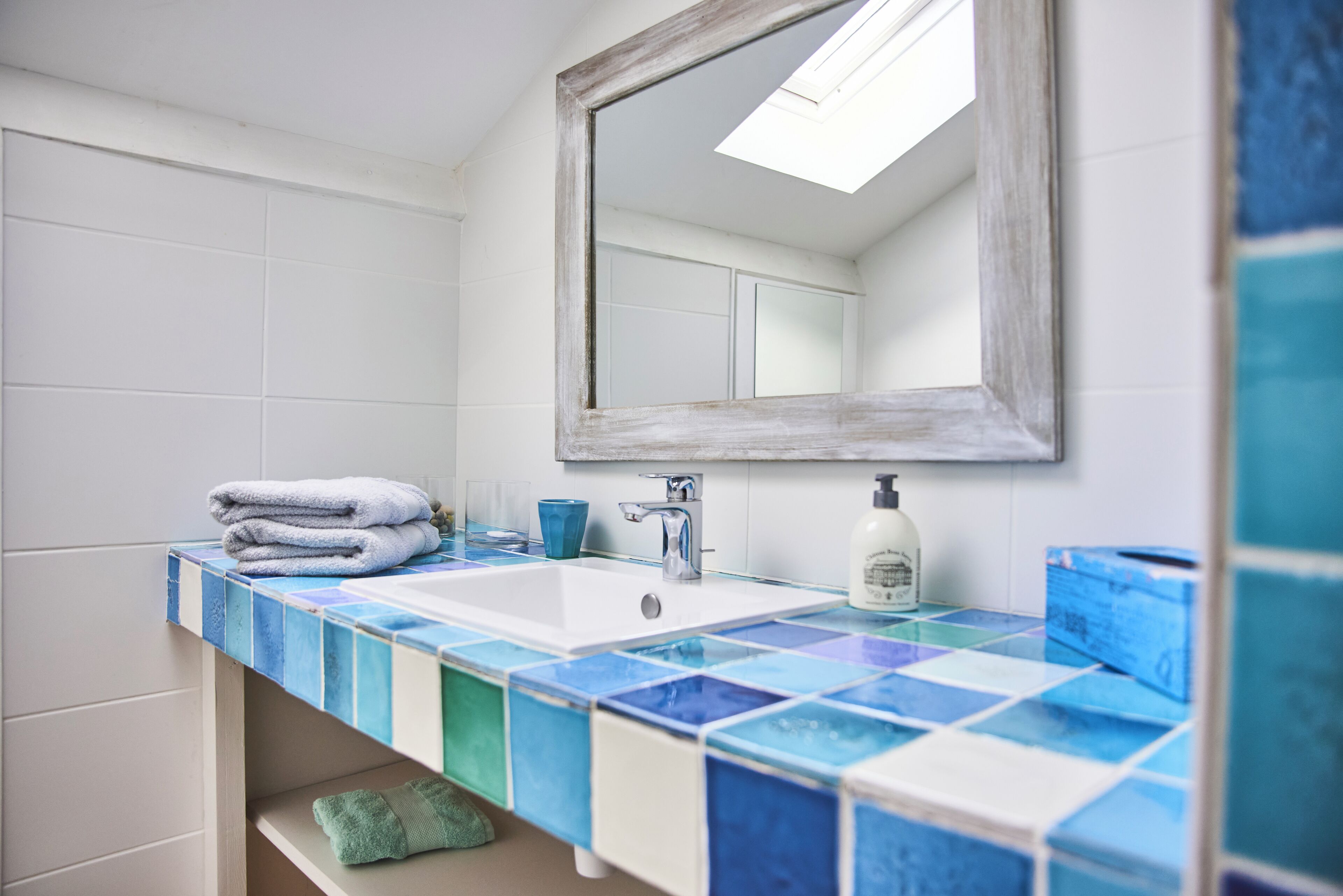 A bathroom with a tiled countertop, a sink, a mirror, and a shelf with towels.